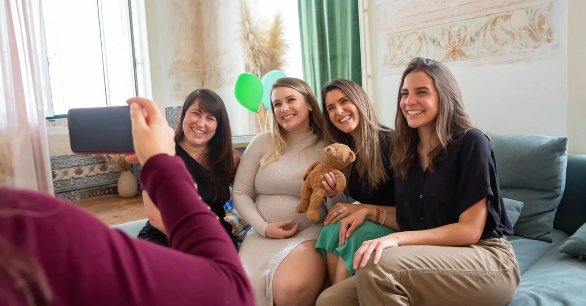 Quatre femmes s'amusant lors d'une joyeuse baby shower à l'intérieur, souriant et posant pour des photos.