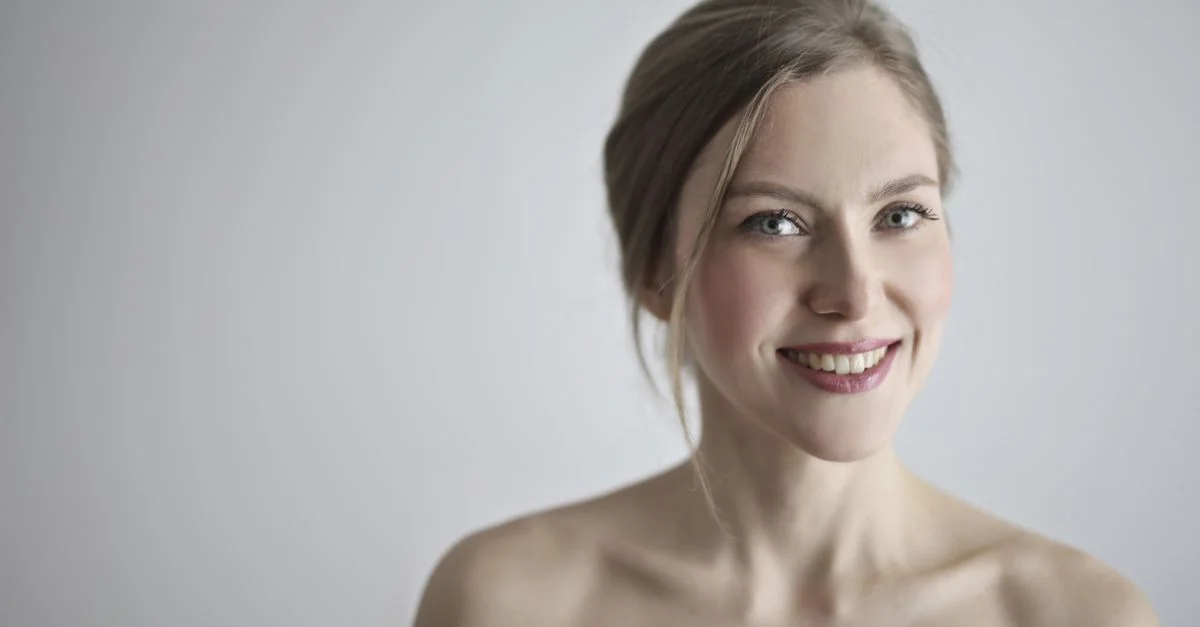 Portrait en gros plan d'une femme blonde souriante sur un fond gris, incarnant élégance et beauté.