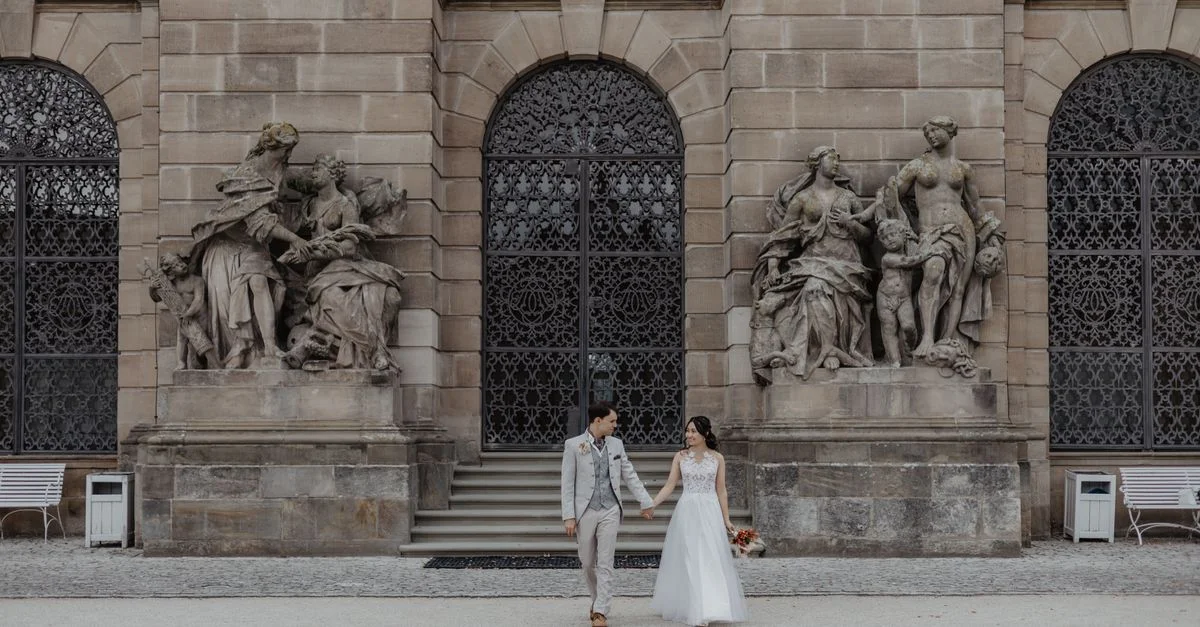 Un couple de jeunes mariés en tenue de cérémonie pose devant un bâtiment historique en Allemagne.