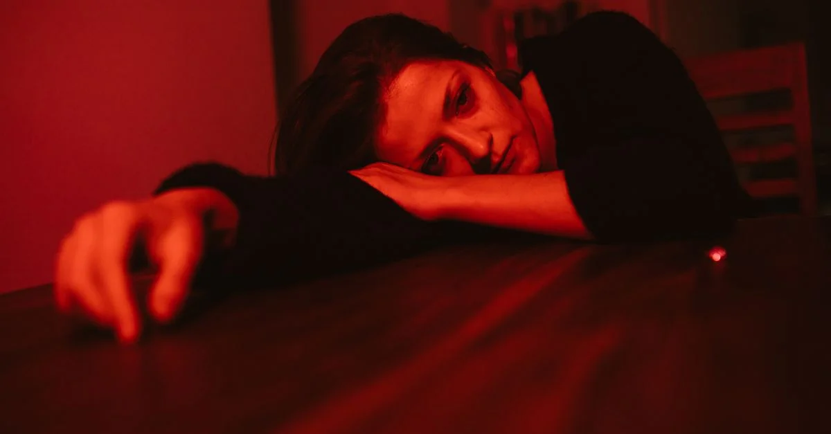 Une femme pensive repose sa tête sur une table, baignée dans une lumière rouge, reflétant l'émotion.